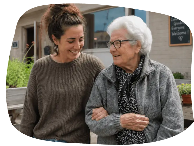 A younger woman and an older woman walk arm in arm outside a community garden, smiling and talking together.
